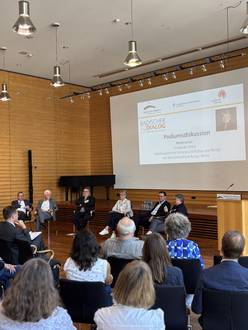 Fünf Personen sitzen am Tisch in der Aula, viele Teilnehmende lauschen der Diskussion.